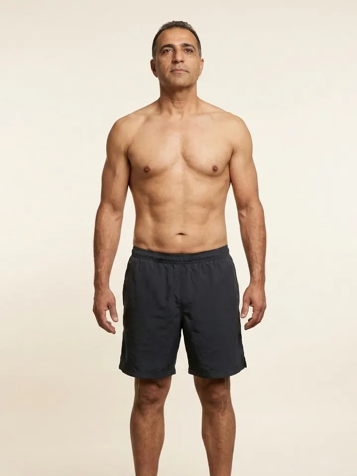 Sample member photo, third visit: the same man, lean and muscular, stands in the same pose in dark shorts against the same plain warm backdrop.