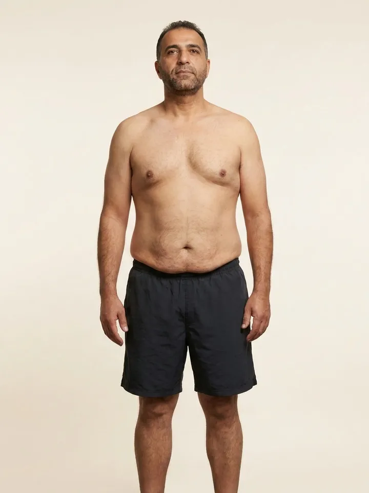 Sample member photo, second visit: the same man, noticeably slimmer, stands in the same pose in dark shorts against the same plain warm backdrop.
