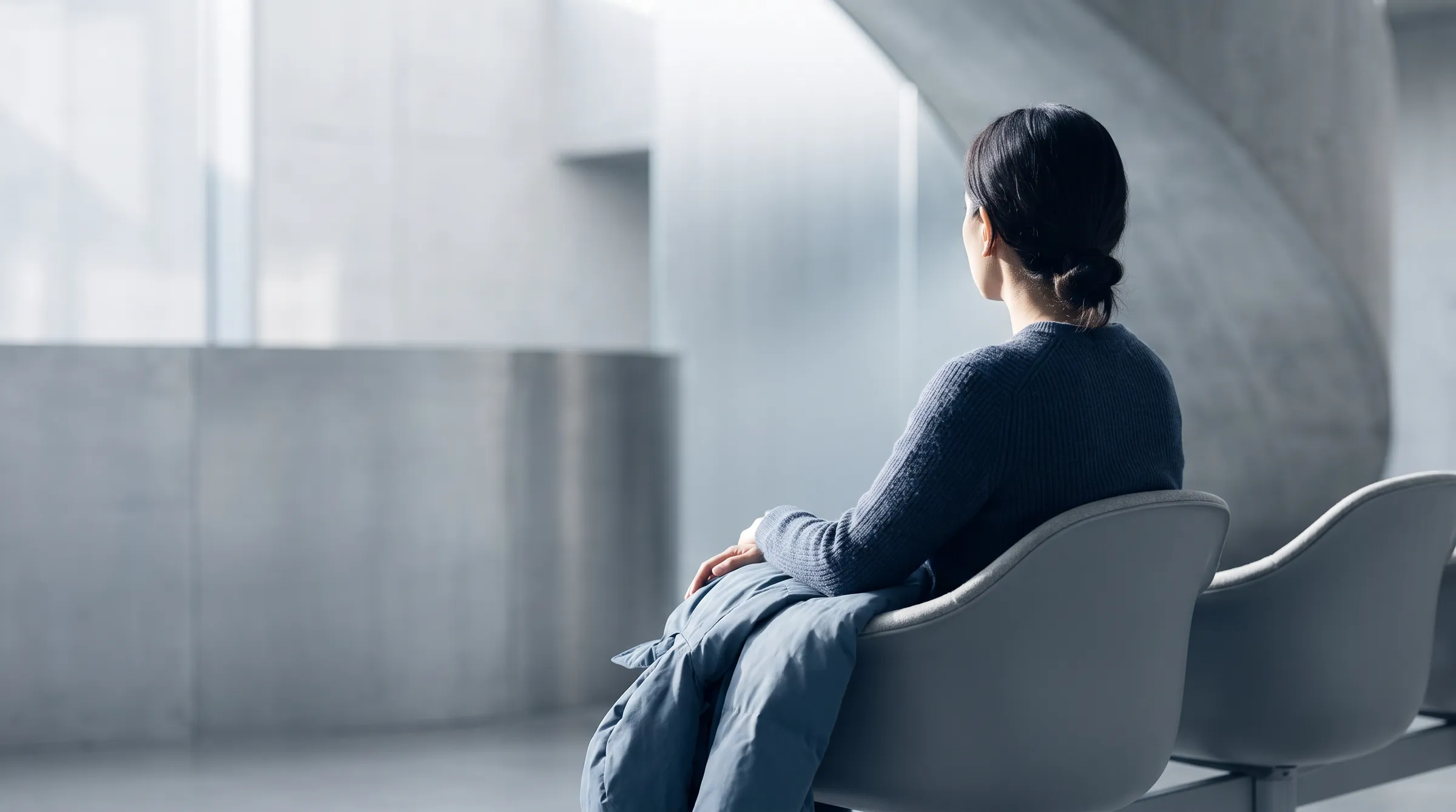 A person sits turned away in a soft chair against pale concrete, morning daylight falling across one shoulder.